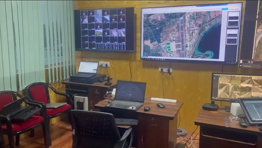 A control room with two wall displays, one showing a grid of camera views and one showing a satellite map, above desks with laptops and monitors.