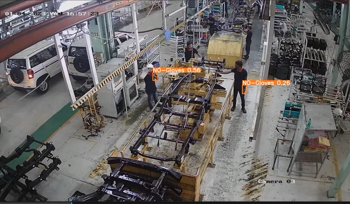 Overhead factory camera view of a vehicle chassis assembly line. Orange boxes mark the bare hands of two workers.